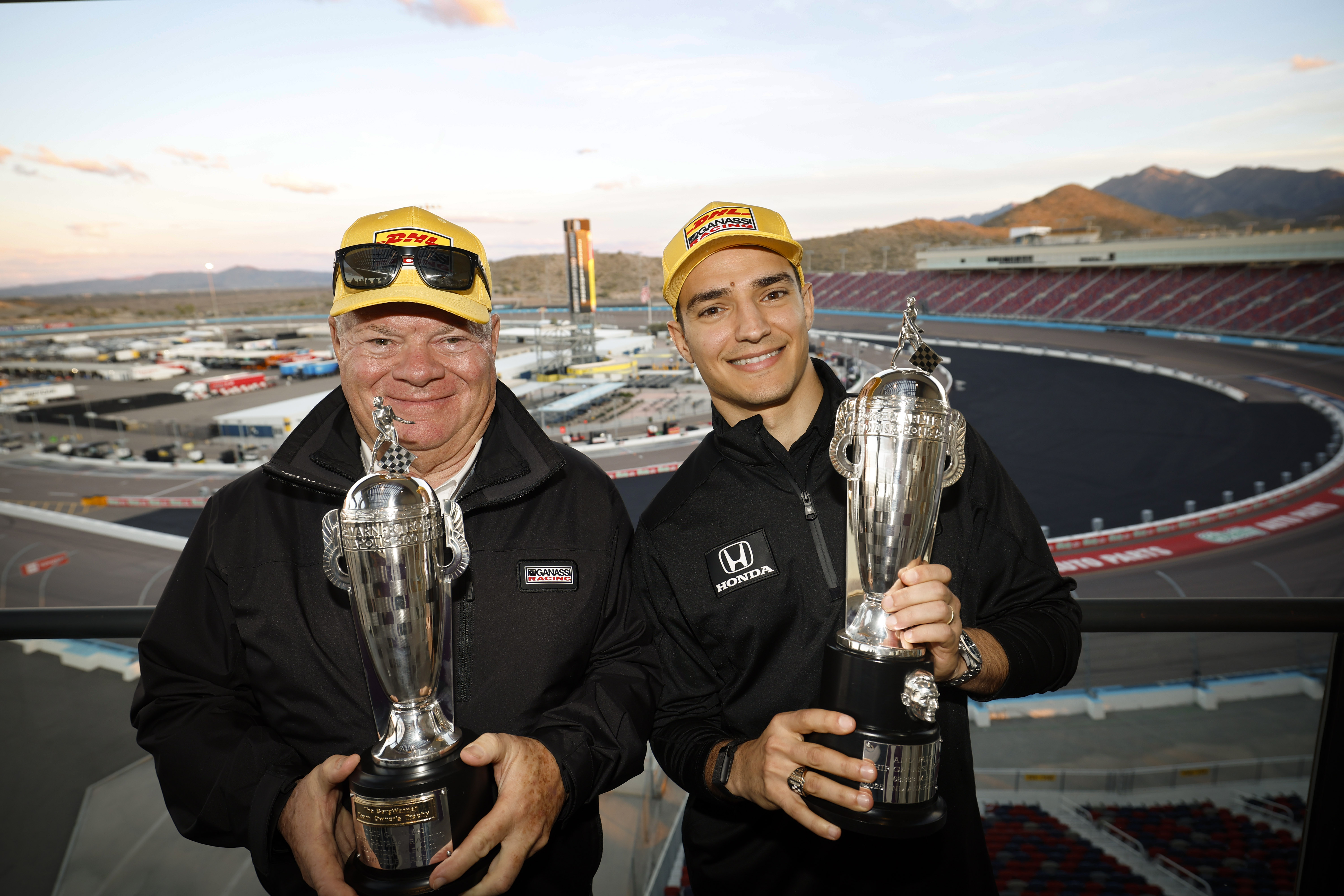 Two men hold trophies and smile in front of race track