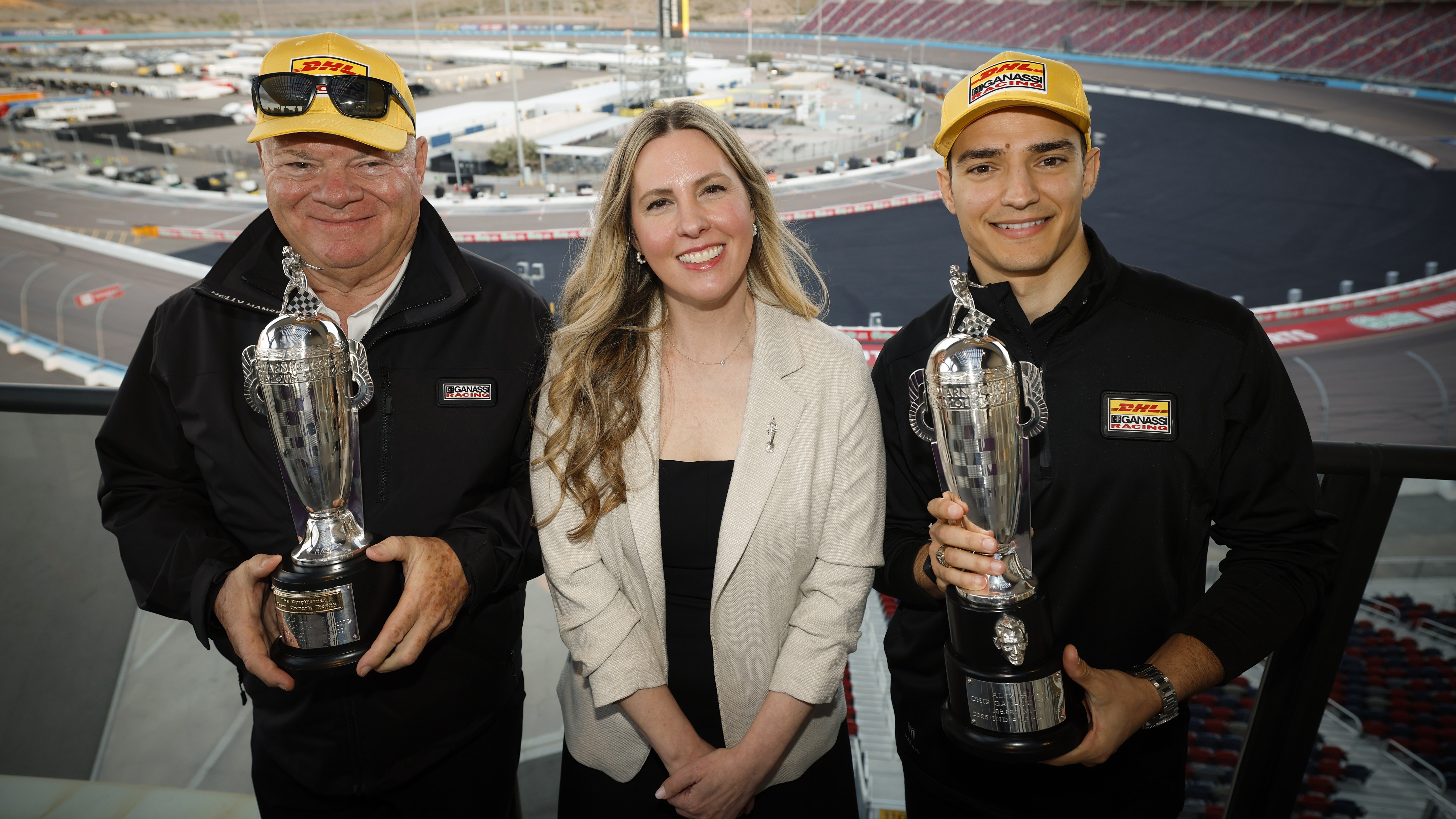 Two men and a woman smile while holding trophies in front of a race track