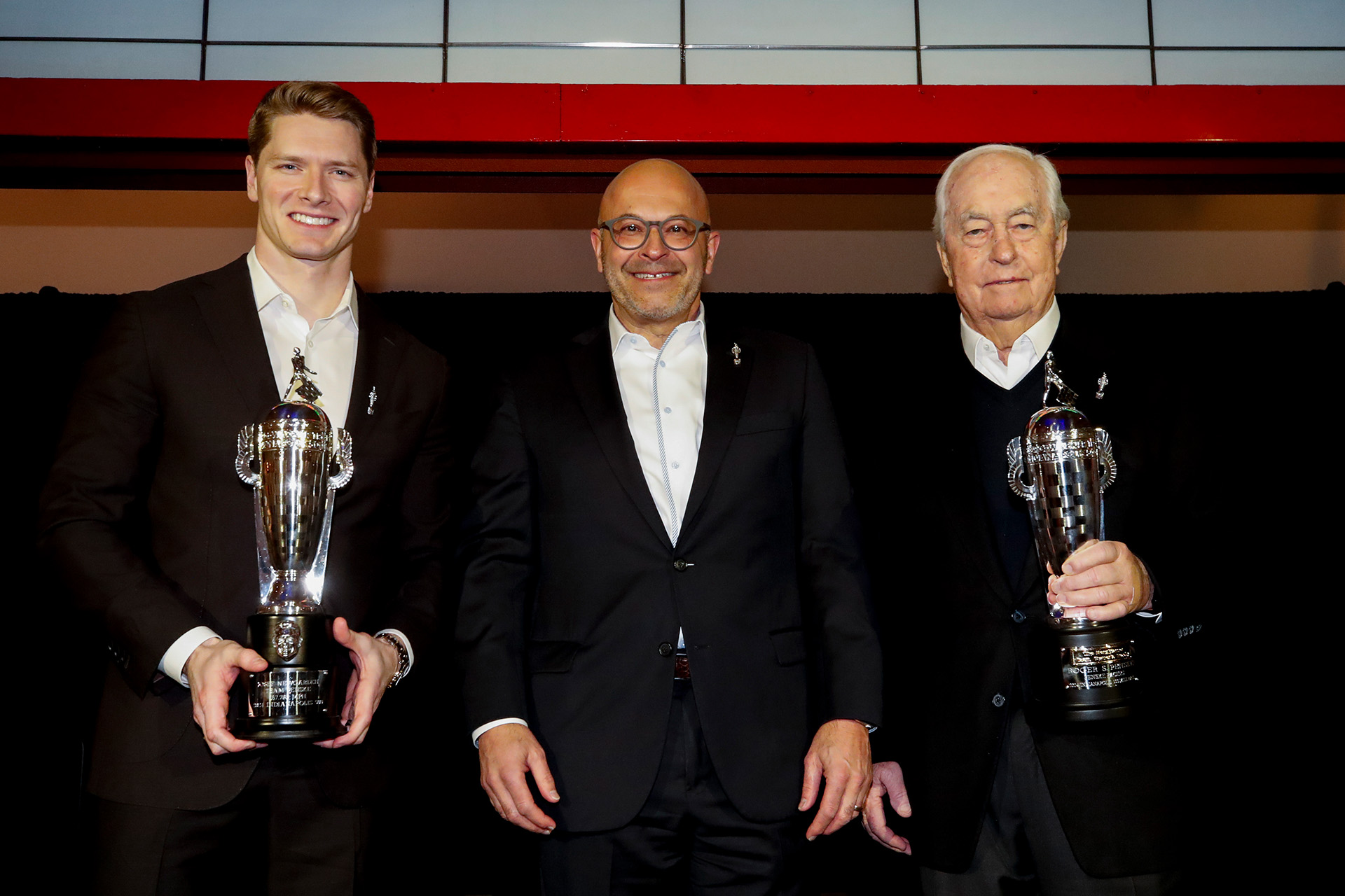 Three men smile on stage with two holding silver trophies
