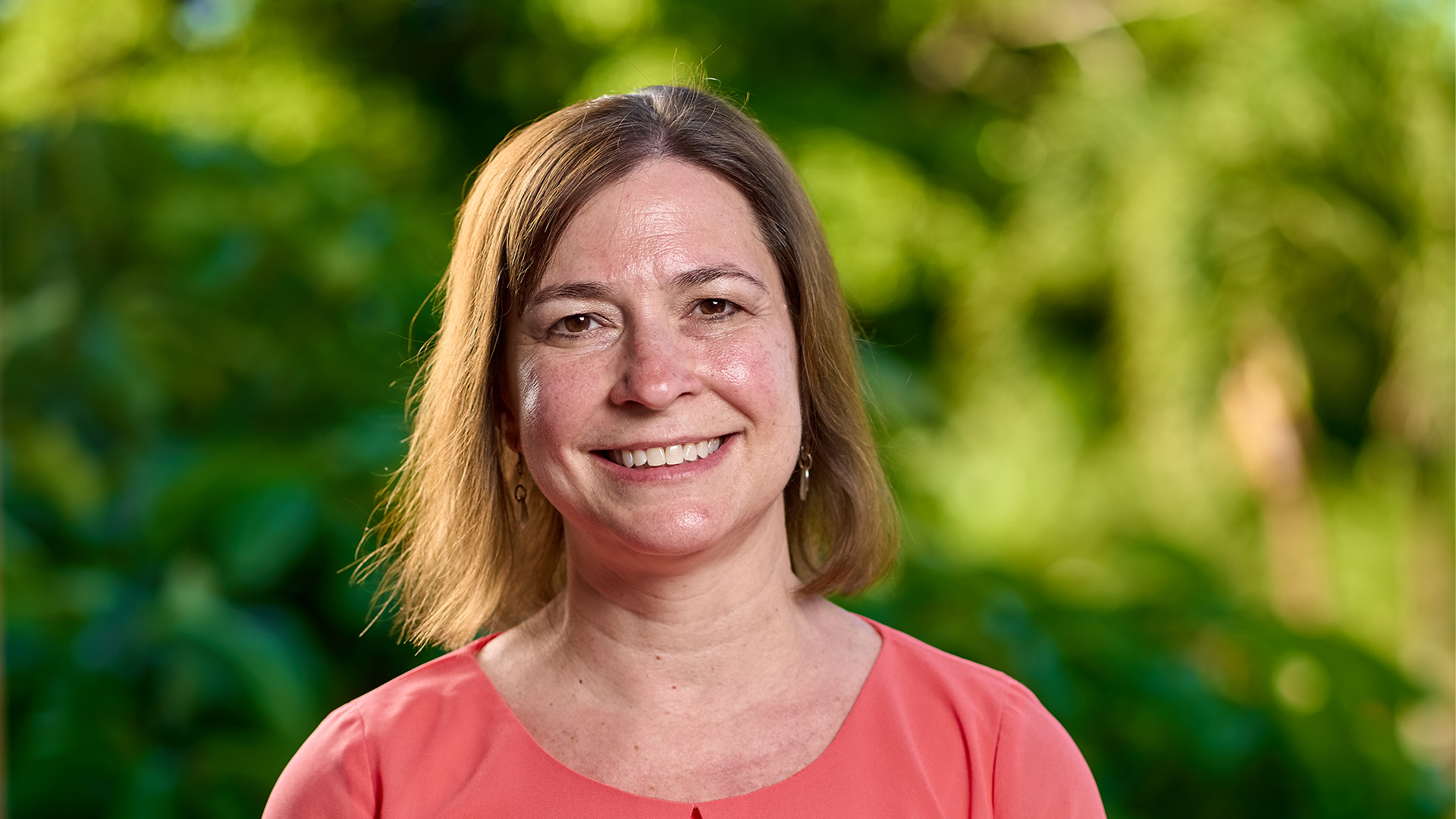 Woman in coral blouse smiles in front of blurred greenery