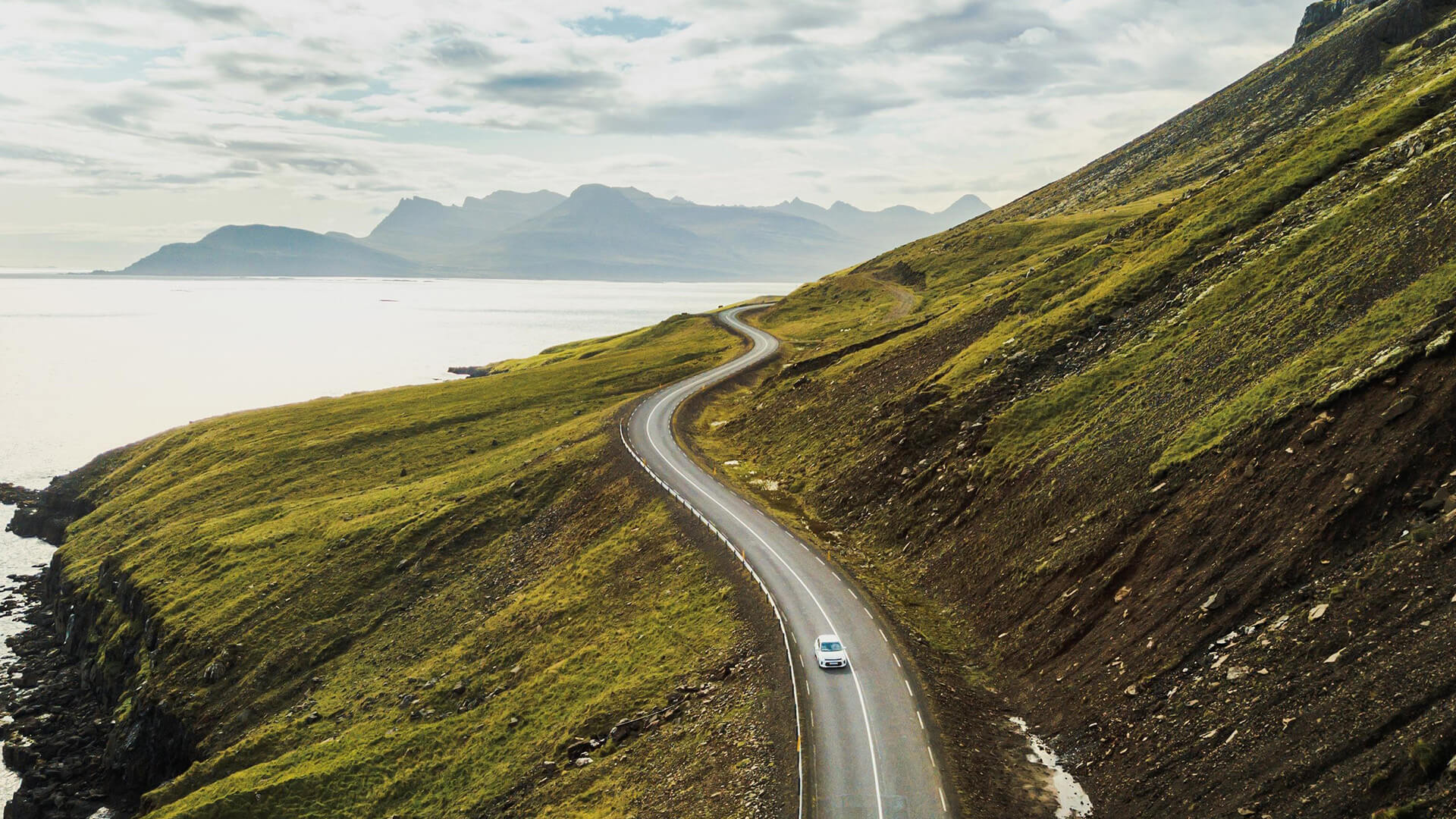 Single car drives down paved road on hilly landscape surrounded by water
